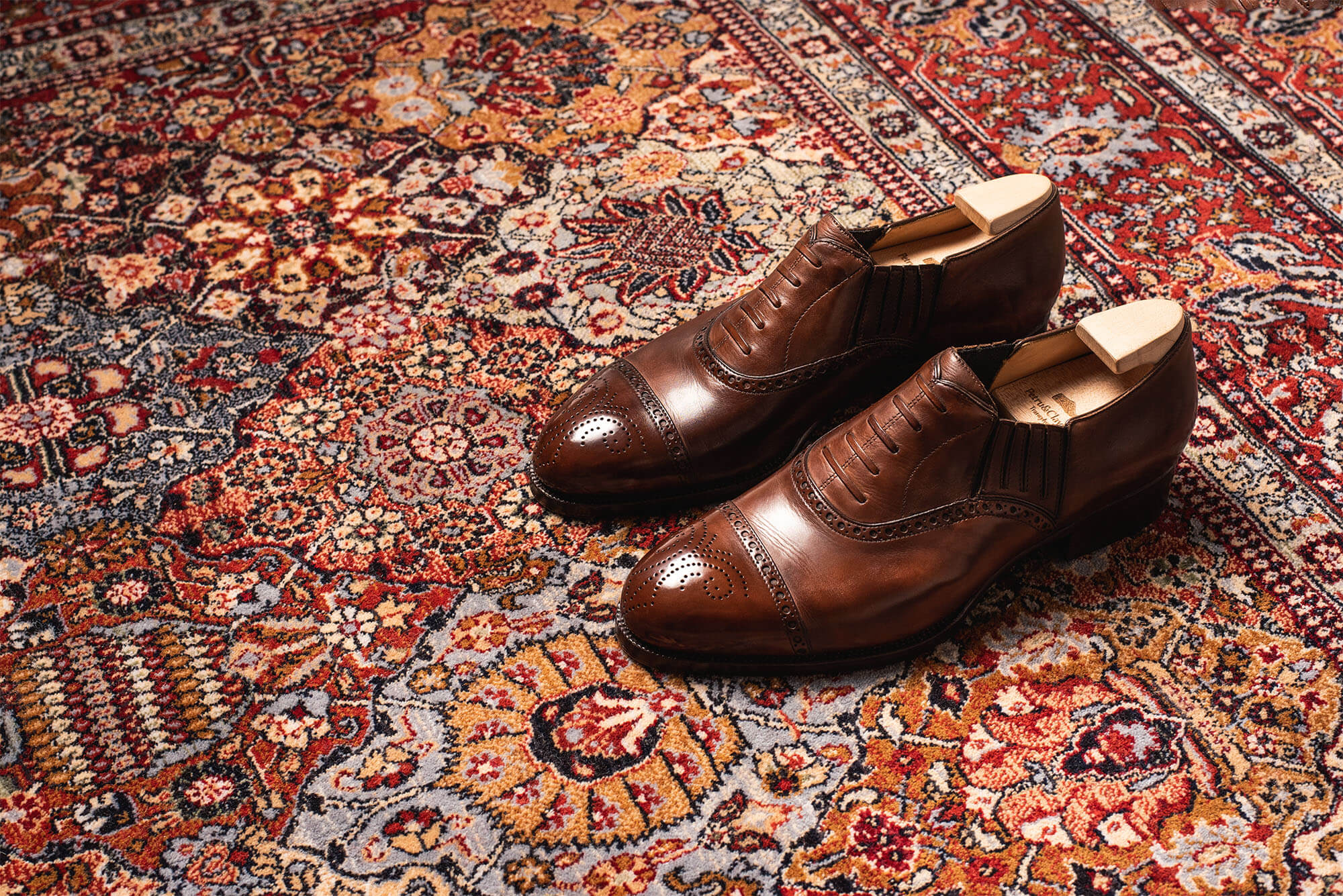 Brown handmade leather shoes with decorative perforations, placed on a patterned rug.