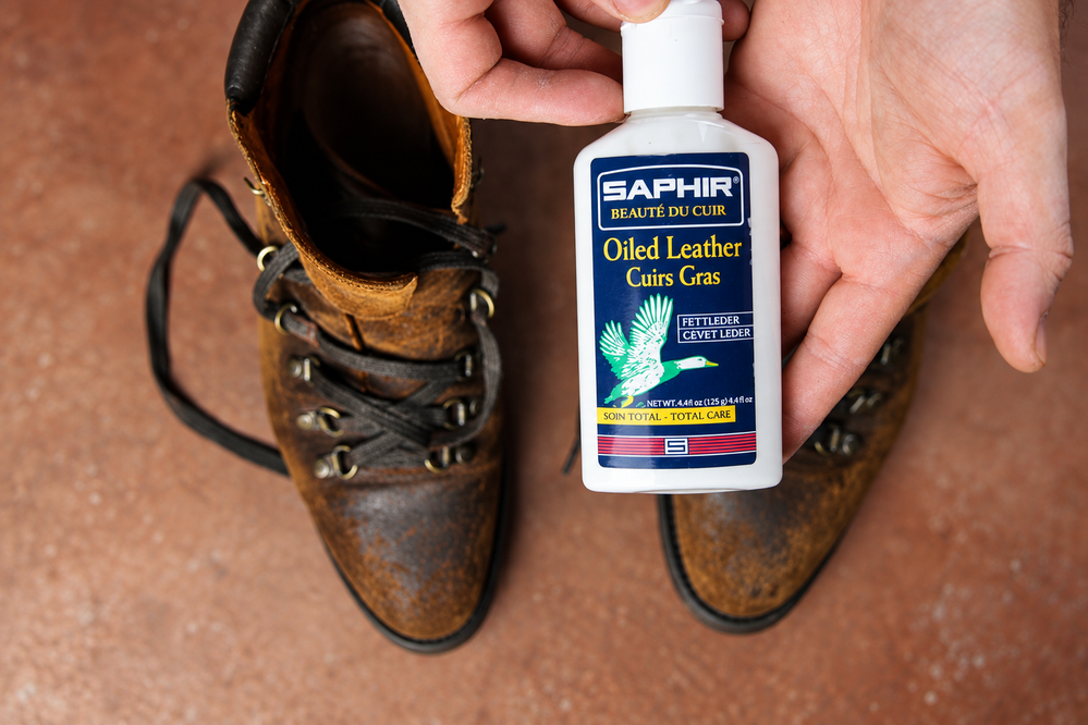 A hand holds a bottle of Saphir Oiled Leather Cream above a pair of worn brown leather boots.