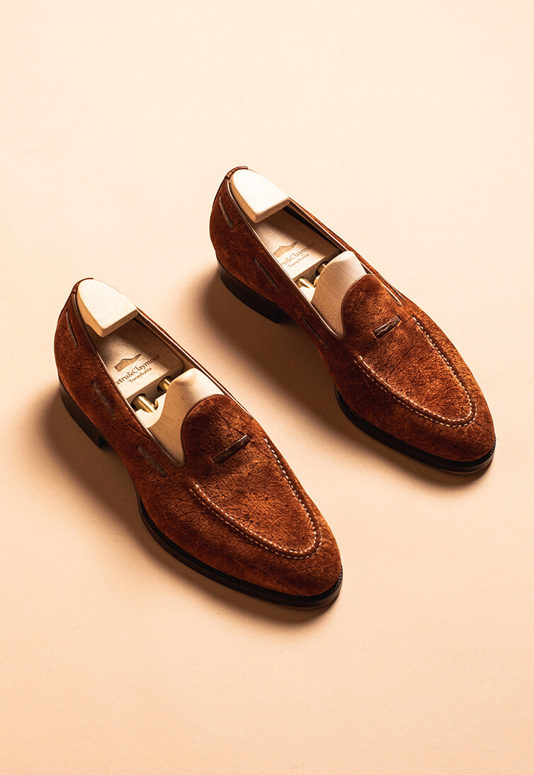 Two handmade brown suede loafers with a stitched design and wooden shoe trees inside.
