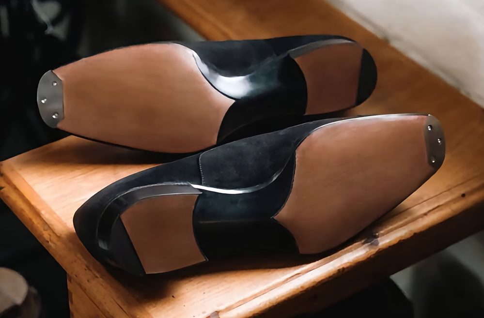 Black suede shoes with smooth leather soles, shown from the bottom.