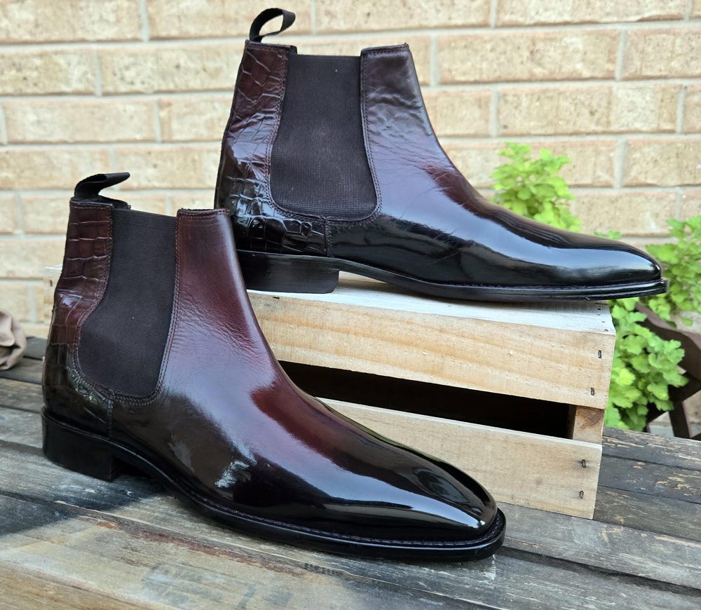 A pair of polished burgundy leather Chelsea boots with black elastic side panels, displayed on a wooden crate.
