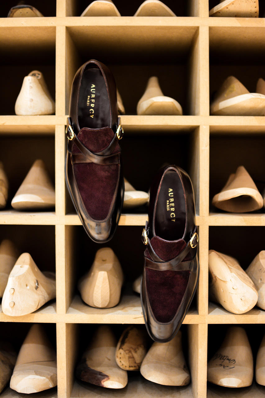 A pair of burgundy suede and leather shoes with gold buckles, displayed on a wooden shelf with shoe molds.