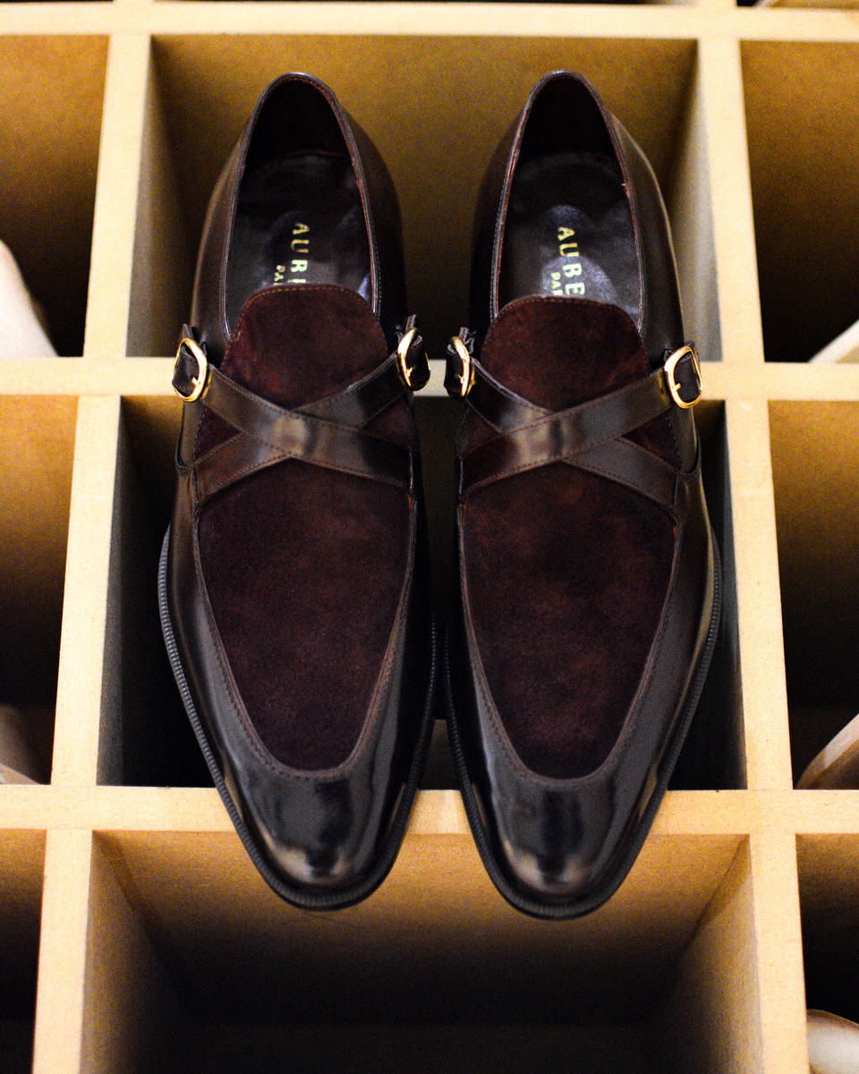 A pair of dark brown suede and polished leather shoes with a cross strap and gold buckle.