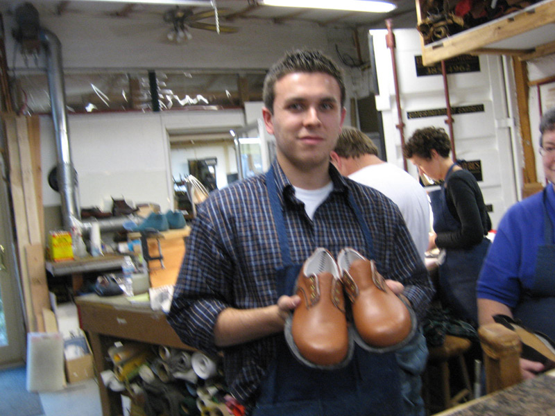 A young man in a checkered shirt holds two brown shoes, showcasing them in a workshop setting.