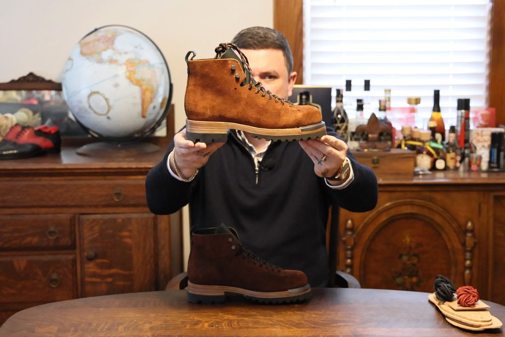 A person holds up a pair of brown suede hiking boots with contrasting black soles, while another similar boot sits on the table.