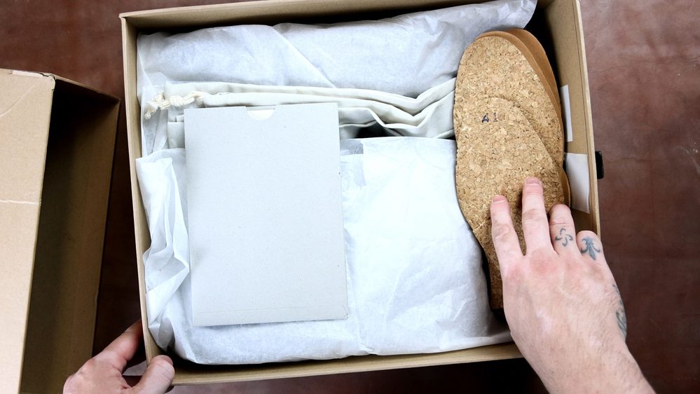 A hand is reaching for a cork sole inside a box containing FEIT wholecut hiking boots, surrounded by tissue paper and a folded cloth.