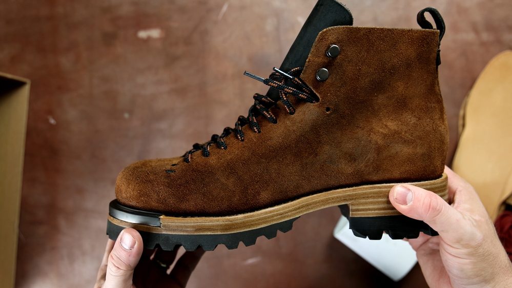A person holds a brown suede FEIT wholecut hiking boot with black laces and a rugged sole.