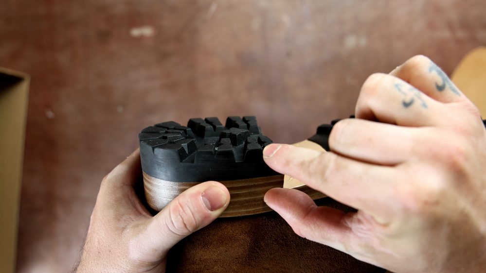 A close-up of hands examining the black rubber sole of FEIT wholecut hiking boots, showcasing its textured tread.
