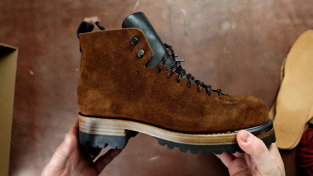 A pair of brown suede FEIT wholecut hiking boots with black leather accents and a rugged sole, being held by hands.