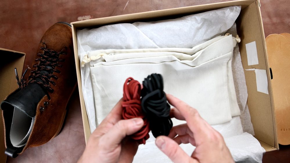 A person holds two sets of laces, one red and one black, next to a brown suede FEIT wholecut hiking boot inside a box.