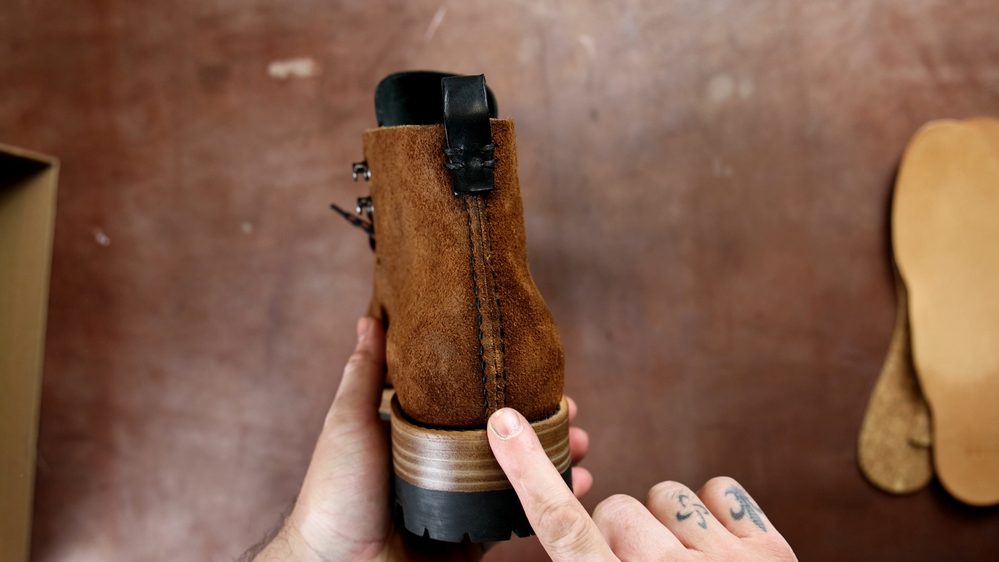 A close-up of the back of brown suede FEIT wholecut hiking boots, featuring a black leather pull tab and a wooden heel.