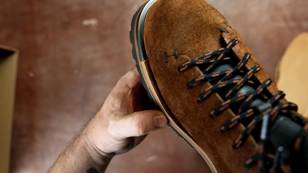 A hand holds a brown suede FEIT wholecut hiking boot with black and orange laces, showcasing its craftsmanship.