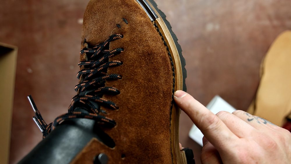 Close-up of FEIT wholecut hiking boots featuring a brown suede upper and black laces, with a finger pointing to the stitching detail.
