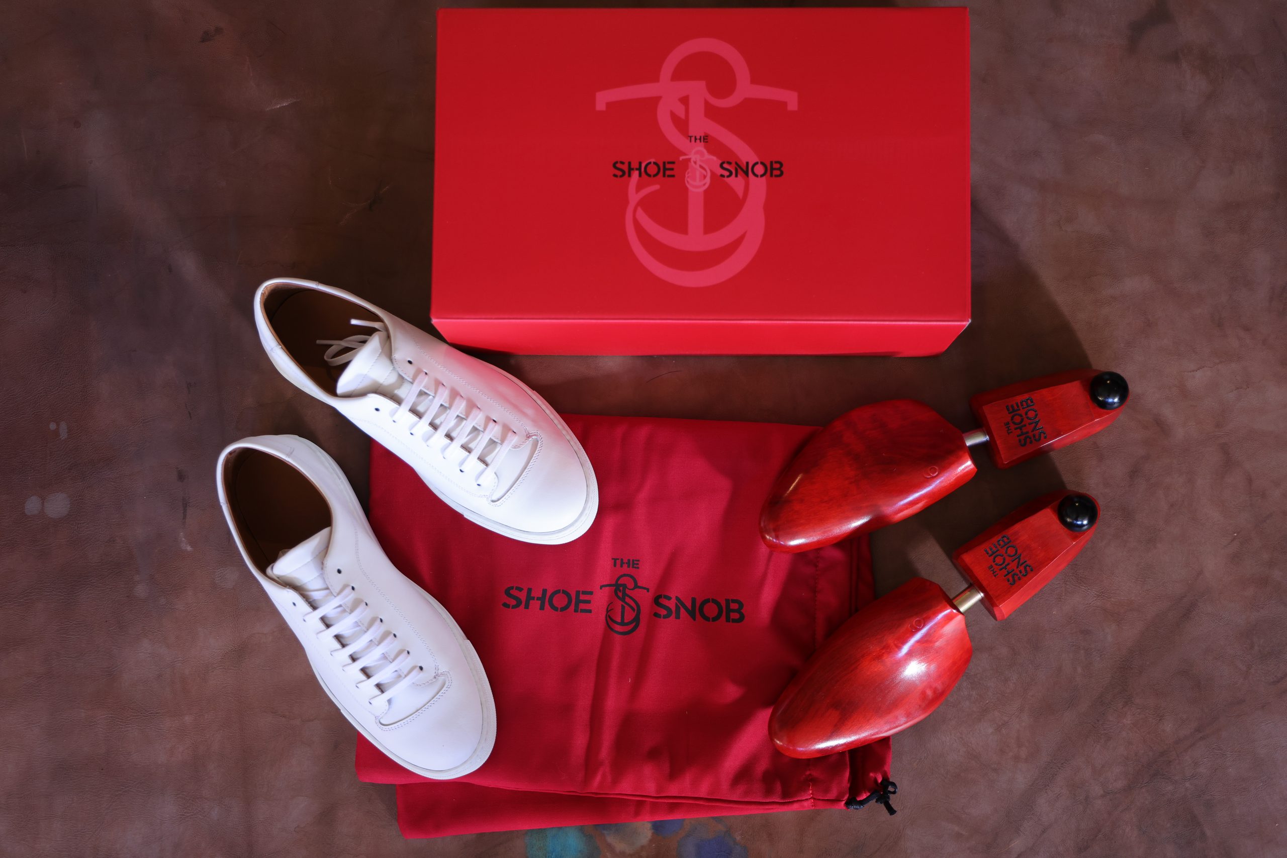 A pair of white shoes, a red shoe bag, and wooden shoe trees are arranged next to a red box labeled "The Shoe Snob," illustrating how to properly store dress shoes.