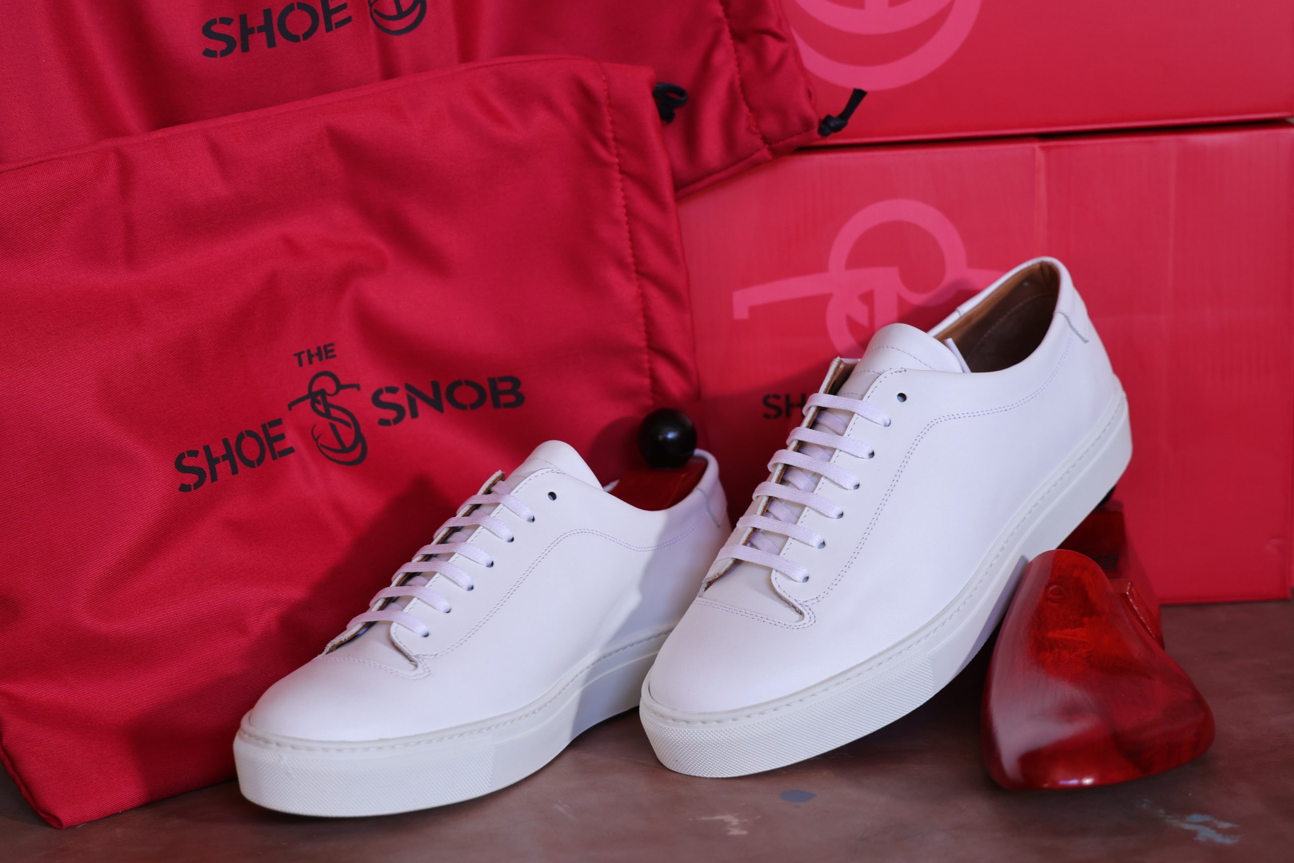 A pair of white dress shoes with a shoe tree and red storage bags labeled "The Shoe Snob," illustrating how to properly store dress shoes.
