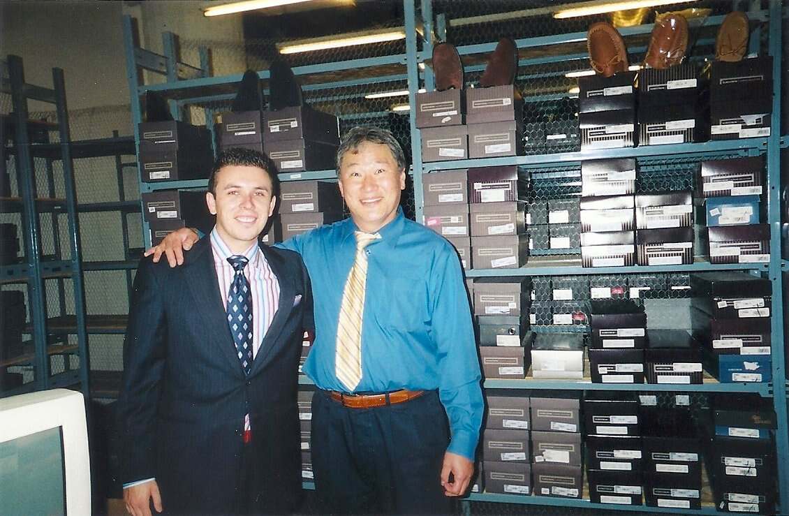 Two men pose together, one wearing a dark suit with a striped shirt and the other in a blue shirt with a yellow tie, in front of shelves filled with shoe boxes.