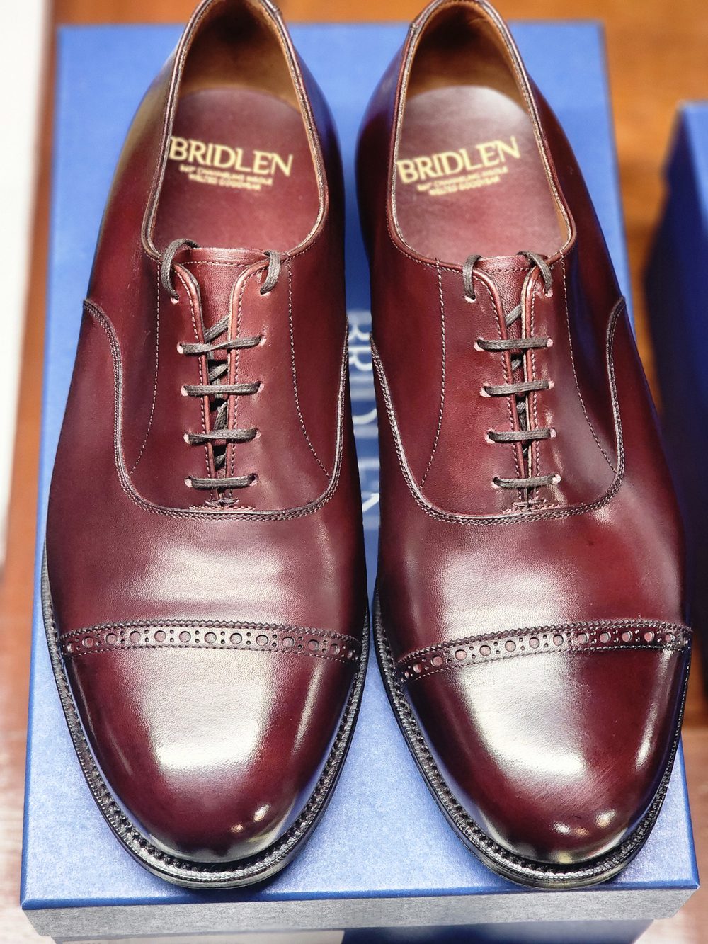 A pair of polished burgundy shoes with decorative broguing and laces, displayed on a blue box.