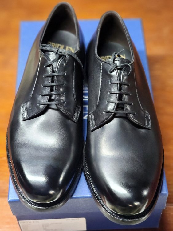 A pair of polished black Oxford shoes with laces, positioned on a blue box, illustrating the Oxford vs wingtip discussion.