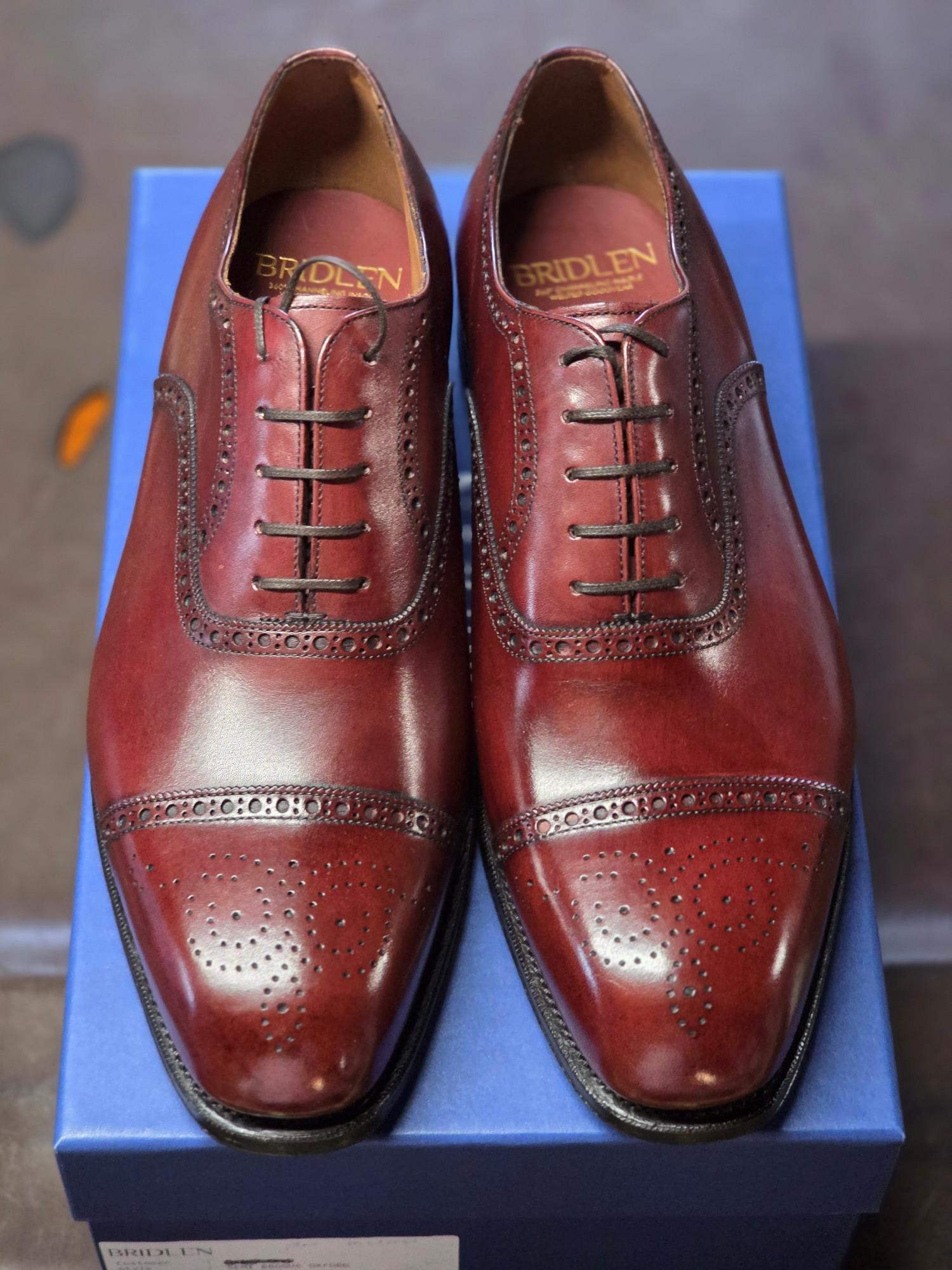 A pair of burgundy leather Oxford shoes with decorative broguing, displayed on a blue box.