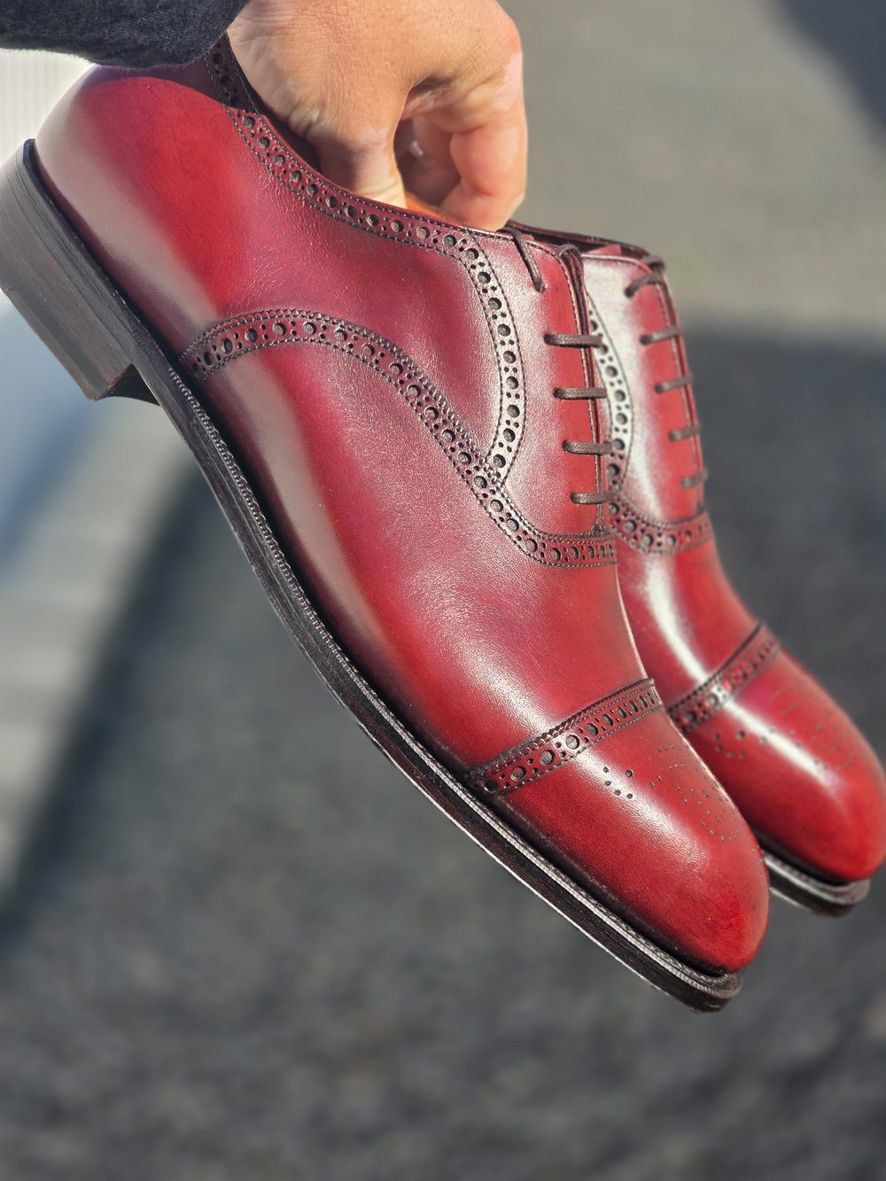 A hand holds a pair of polished burgundy shoes featuring decorative perforations and a classic lace-up design.