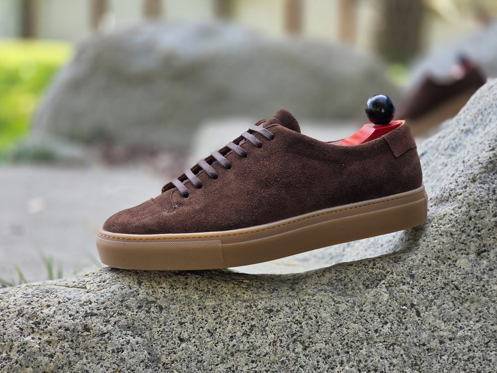 Brown suede sneaker with a gum sole, displayed on a rock.