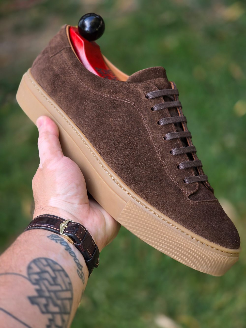 A hand holds a brown suede sneaker with a gum sole and brown laces.