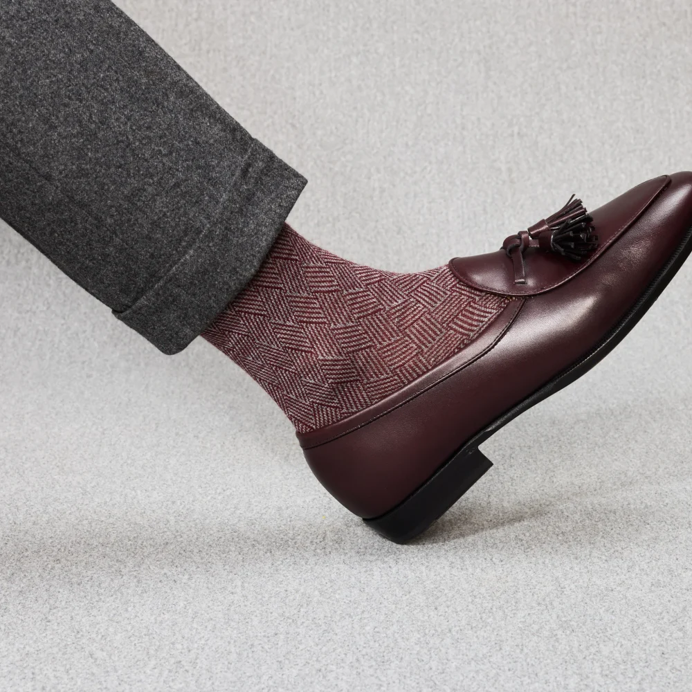 A foot in a burgundy patterned over-the-calf sock paired with a dark leather loafer and gray trousers, illustrating the comparison of over the calf vs mid calf socks.
