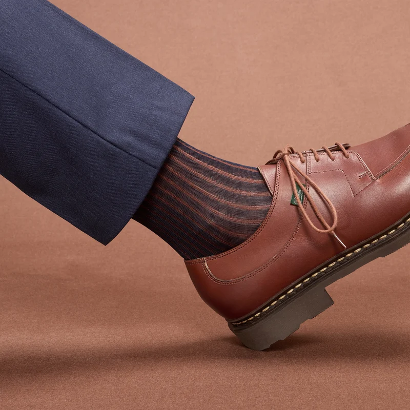 A brown leather shoe and a leg wearing dark striped socks are shown, illustrating the style differences in over the calf vs mid calf socks.