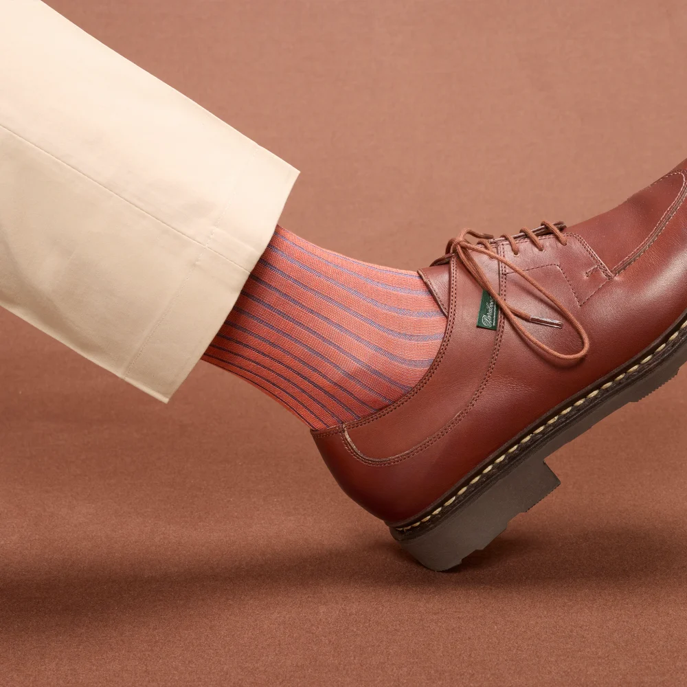 A leg wearing striped orange and blue socks is paired with a brown leather shoe, illustrating the comparison of over the calf vs mid calf socks.