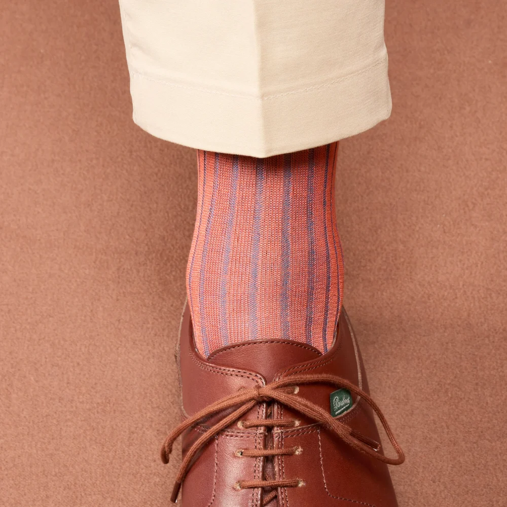 A close-up of a foot wearing striped, over-the-calf socks in shades of orange and blue, paired with brown shoes and beige pants, illustrating the comparison of over the calf vs mid calf socks.