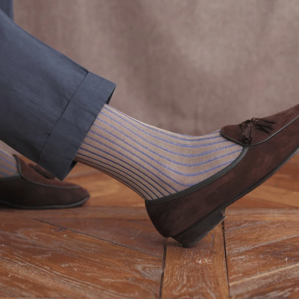 A close-up of a foot wearing striped over-the-calf socks in light blue and beige, paired with a brown suede shoe and navy trousers.