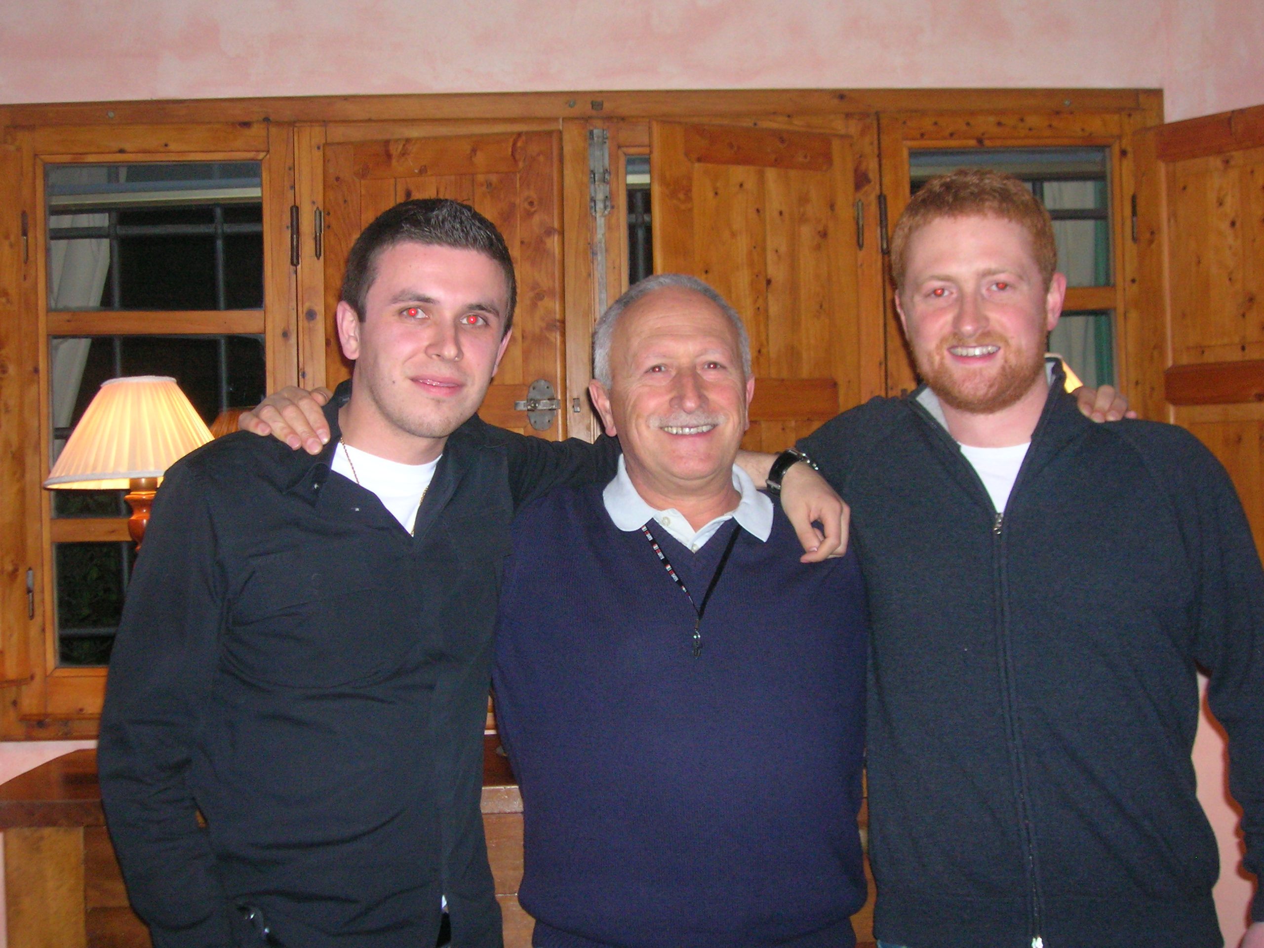 Three men stand closely together, with the central man wearing a navy sweater and the others in black and gray clothing.