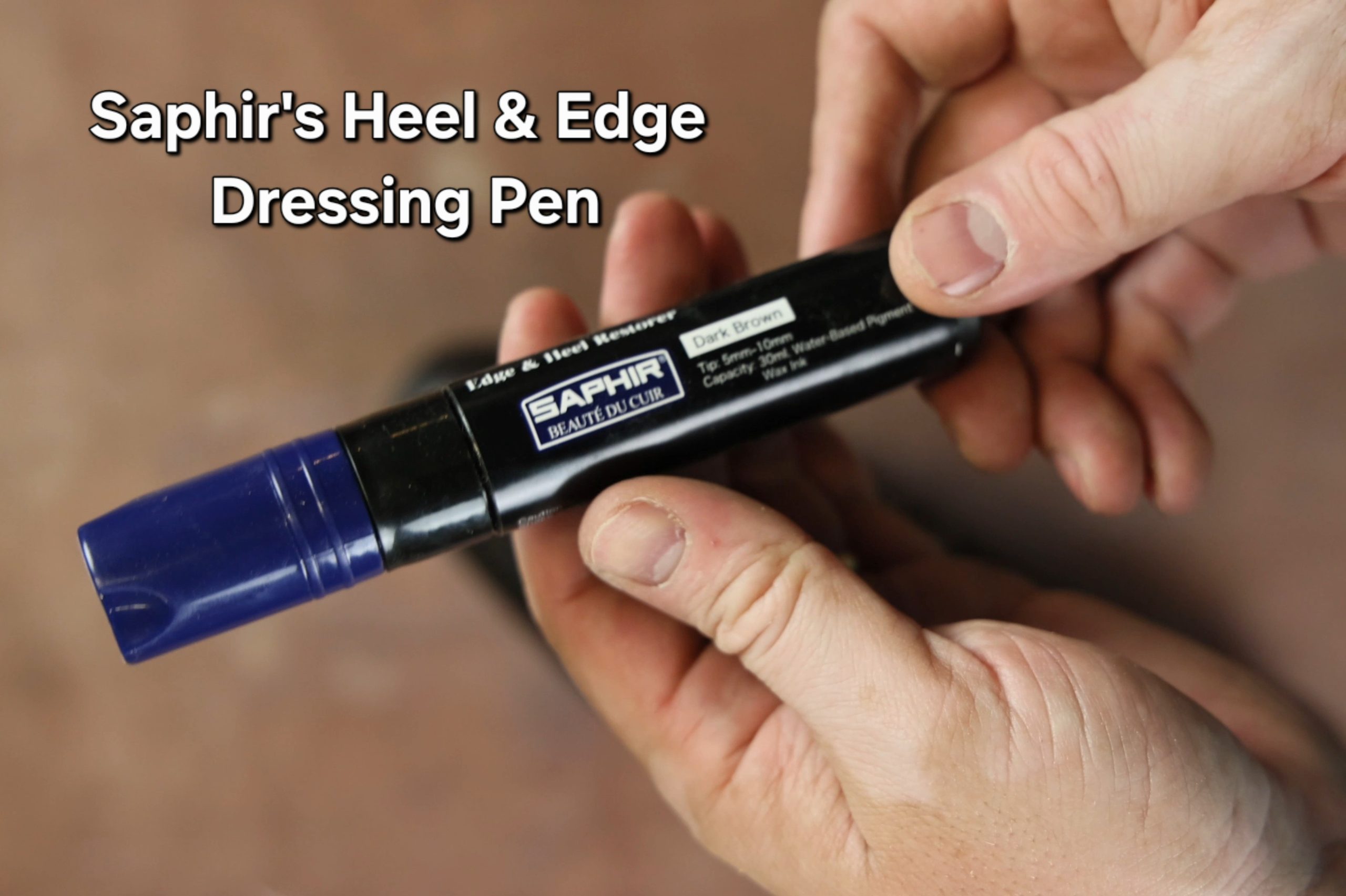 A person holds a black Saphir Heel & Edge Dressing Pen with a blue cap, labeled "Dark Brown."