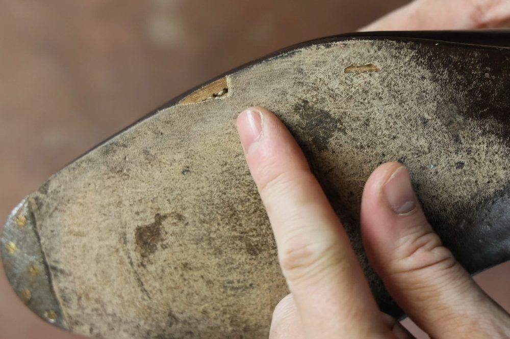 A hand points to a damaged area on a worn leather shoe sole.