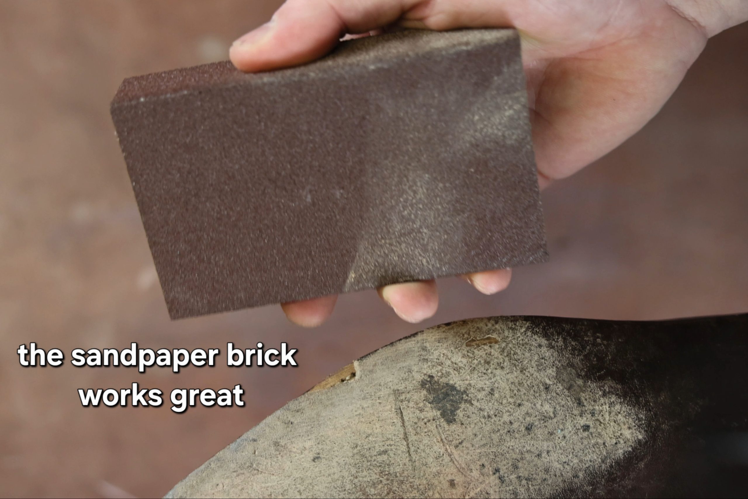 A hand holds a brown sandpaper brick above a worn leather sole.