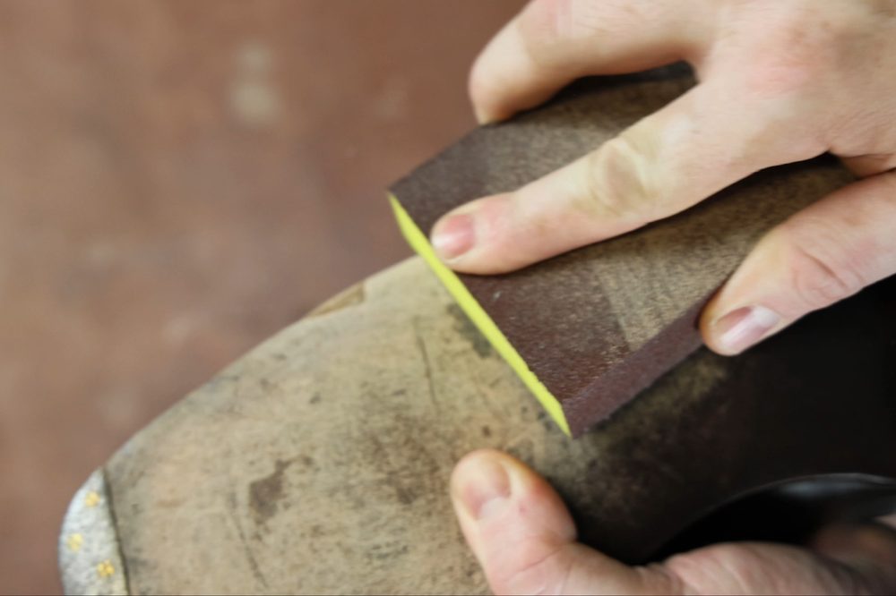 A hand holds a brown sanding block with a yellow edge, smoothing a worn leather sole.