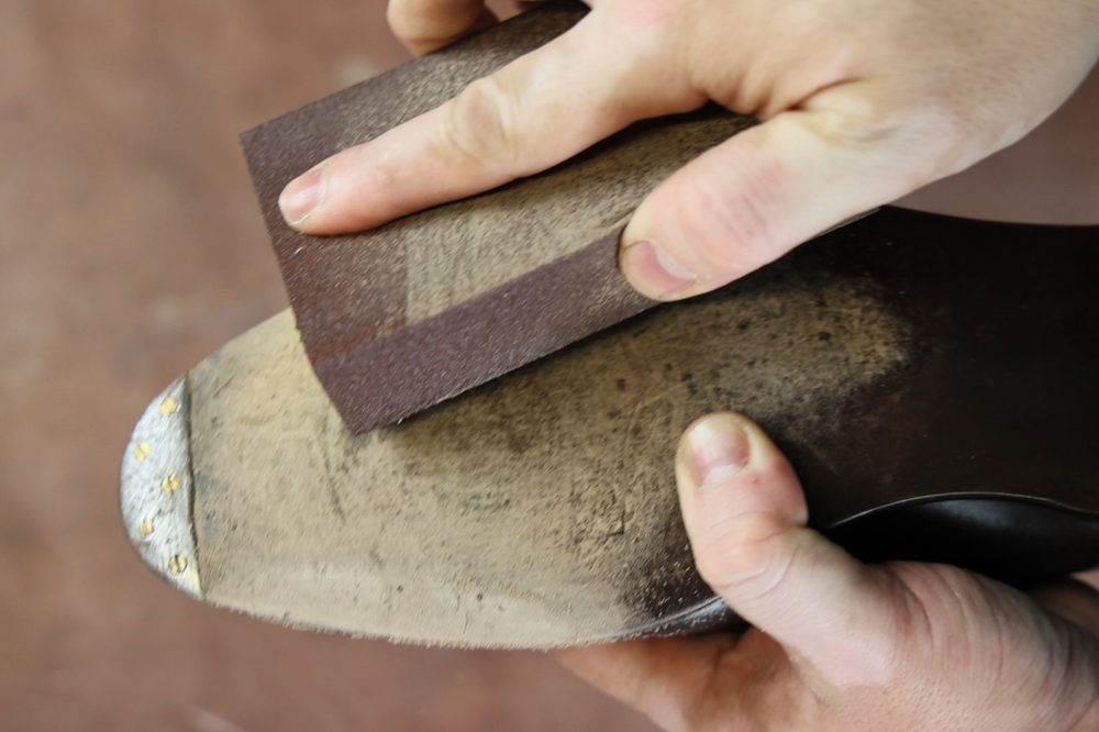 A person sands a damaged closed-channel leather sole with a piece of sandpaper.
