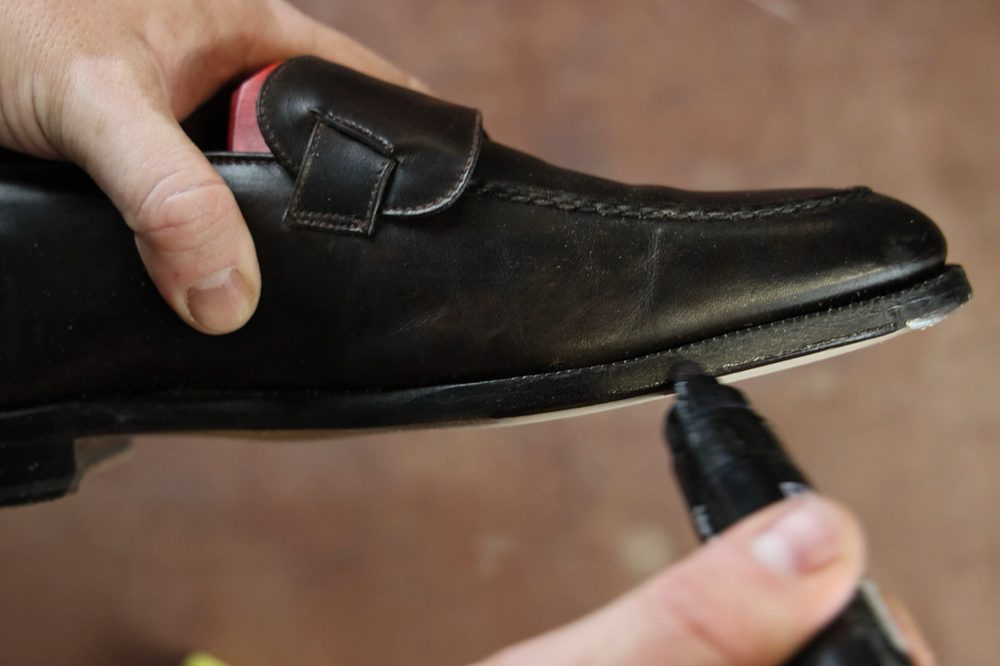 A person is holding a black leather shoe while using a black marker to touch up the sole.