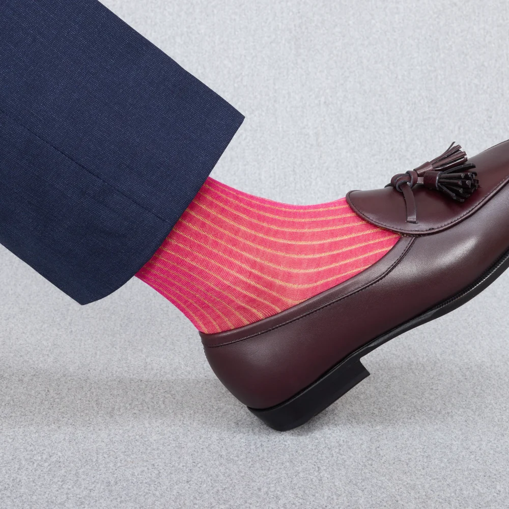 A foot in a dark suit and a brown loafer shows bright pink striped socks, illustrating the style contrast in over the calf vs mid calf socks.