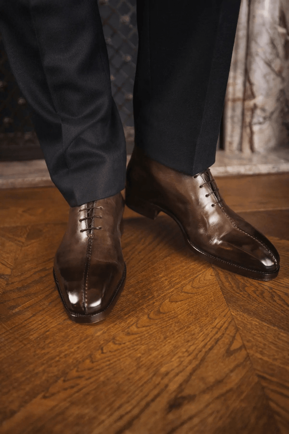 A close-up of polished brown leather dress shoes with decorative lacing, worn with dark trousers.