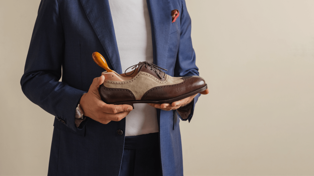 A person in a blue suit holds a two-tone brown and beige dress shoe with a shoehorn.