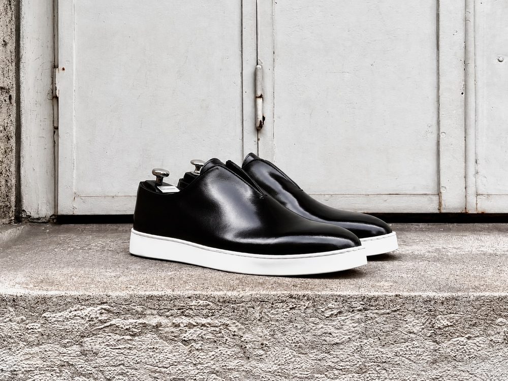 A pair of sleek black leather sneakers with a white sole, featuring a slip-on design.