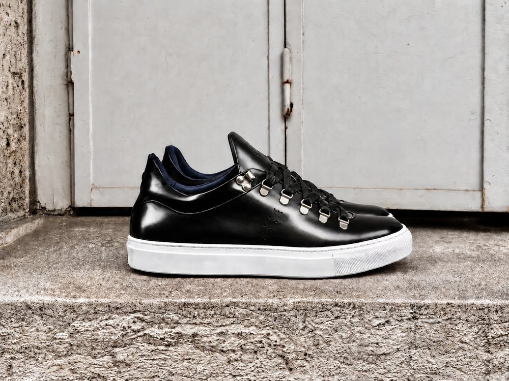 A black leather sneaker with silver eyelets and a white sole, featuring a navy blue interior.