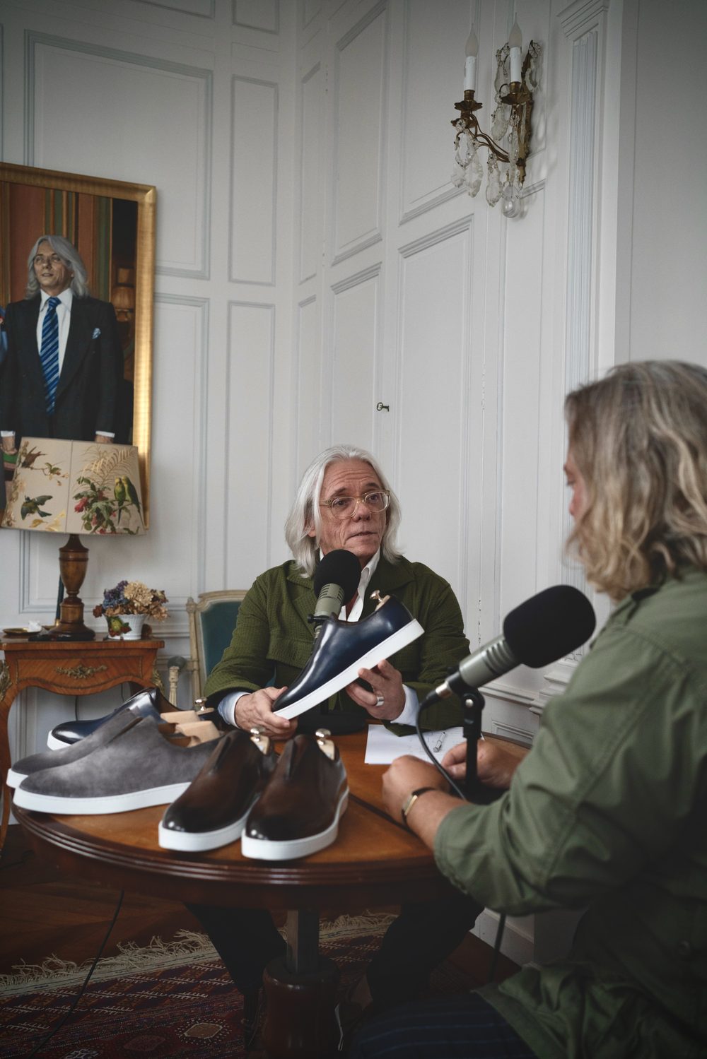 Two men are seated at a table discussing sartorial sneakers, with one man holding a navy sneaker and several other shoes in shades of gray and brown on the table.