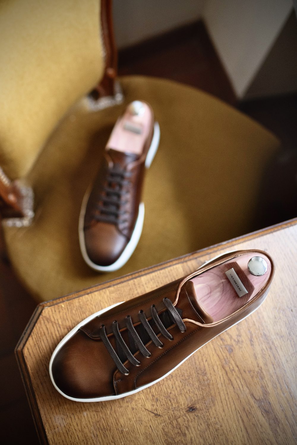 A pair of brown leather sneakers with dark laces and a white sole, placed on a wooden surface.