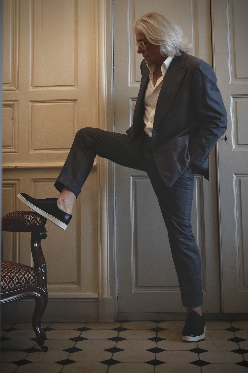 A man in a tailored suit and white shirt raises one leg to showcase a black sartorial sneaker.