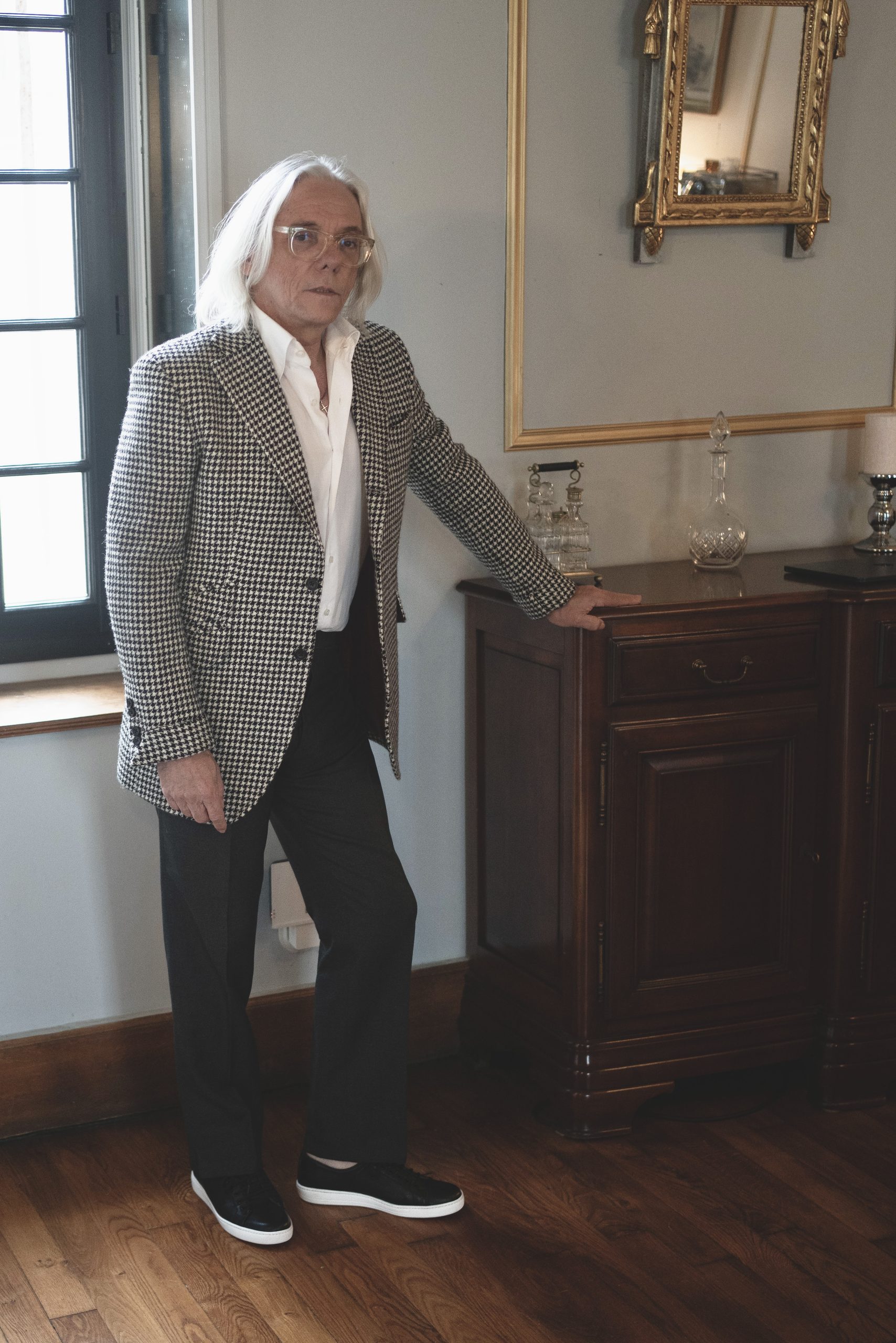 A man with long gray hair and glasses stands beside a wooden cabinet, wearing a houndstooth blazer, white shirt, dark pants, and black sneakers.