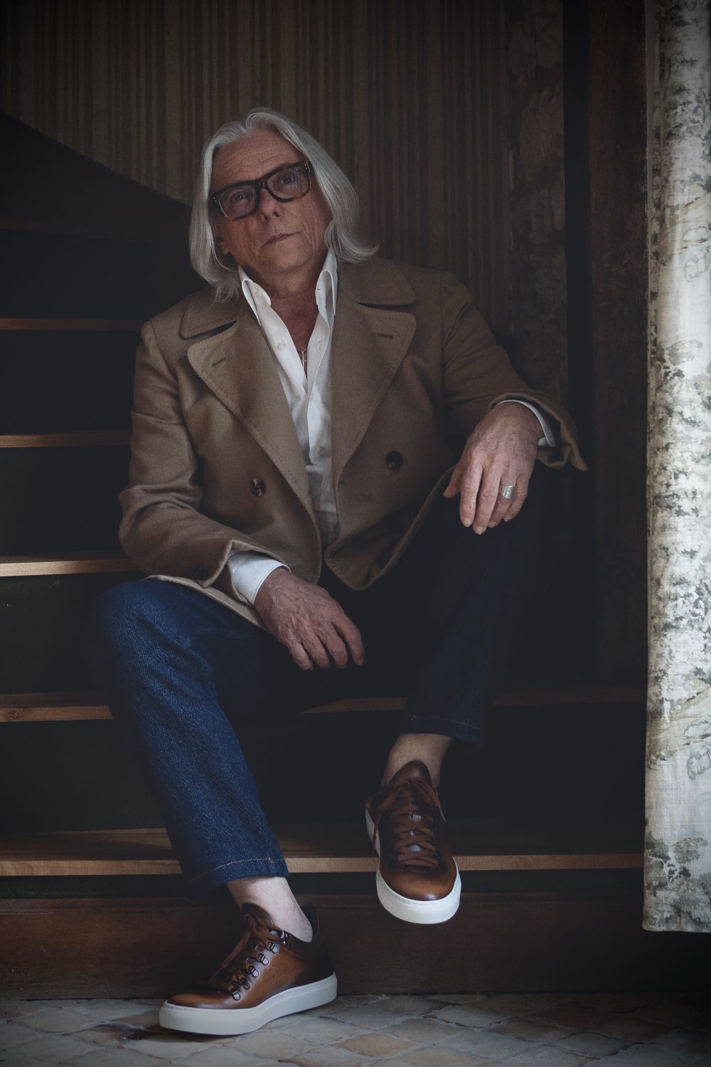 A man with long gray hair and glasses sits on stairs, wearing a beige coat and brown sartorial sneakers.
