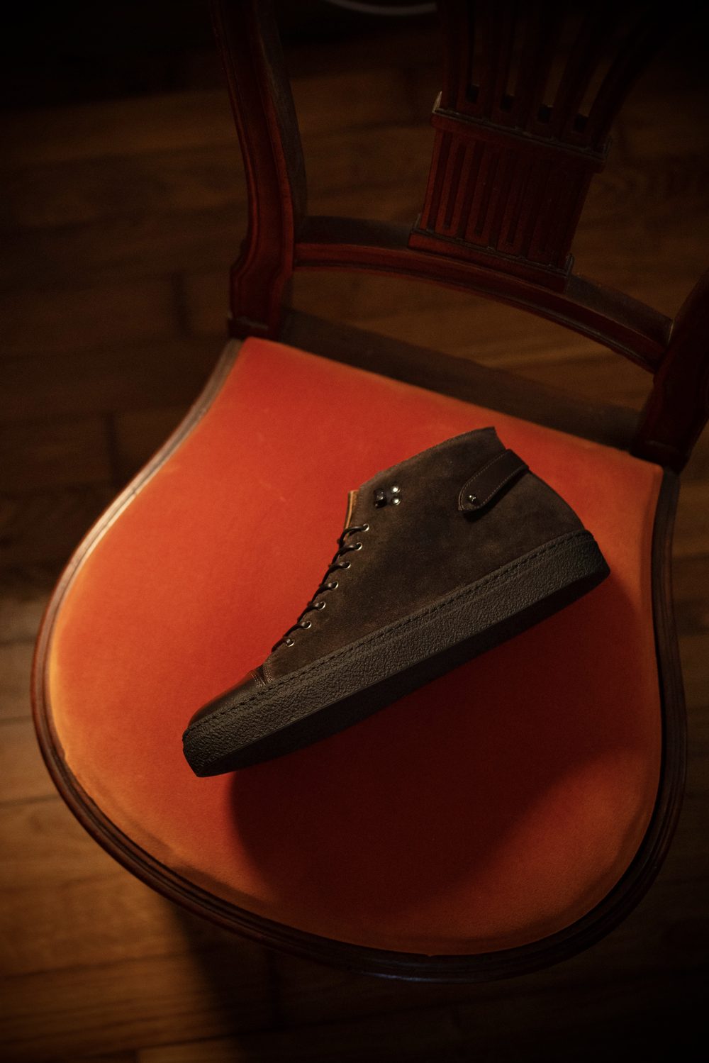A brown suede high-top sneaker with black rubber soles is placed on an orange velvet chair.