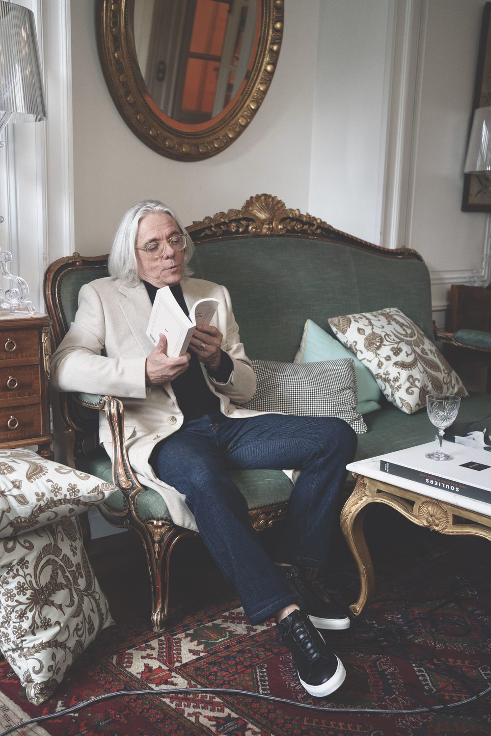 An older man with long gray hair sits on a green upholstered sofa, wearing a beige blazer and reading a book.